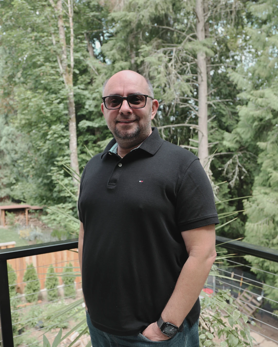 Adrian, systems analyst, standing outdoors on a balcony with trees in the background.