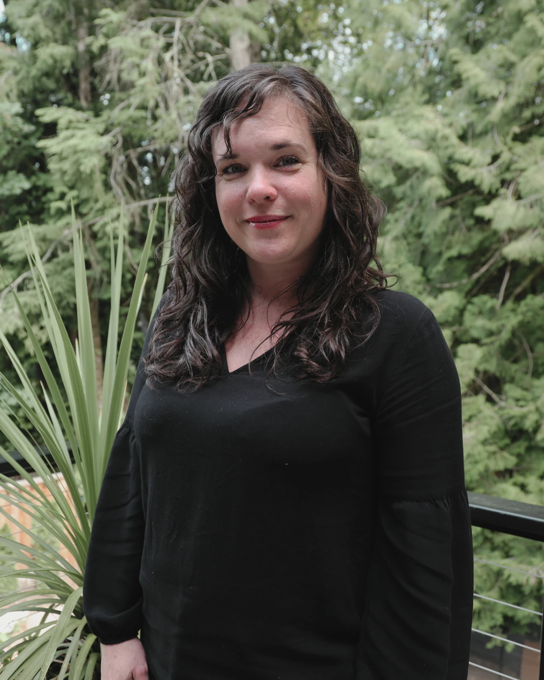 Nikki, senior designer, standing outdoors on a balcony with trees and plants in the background.