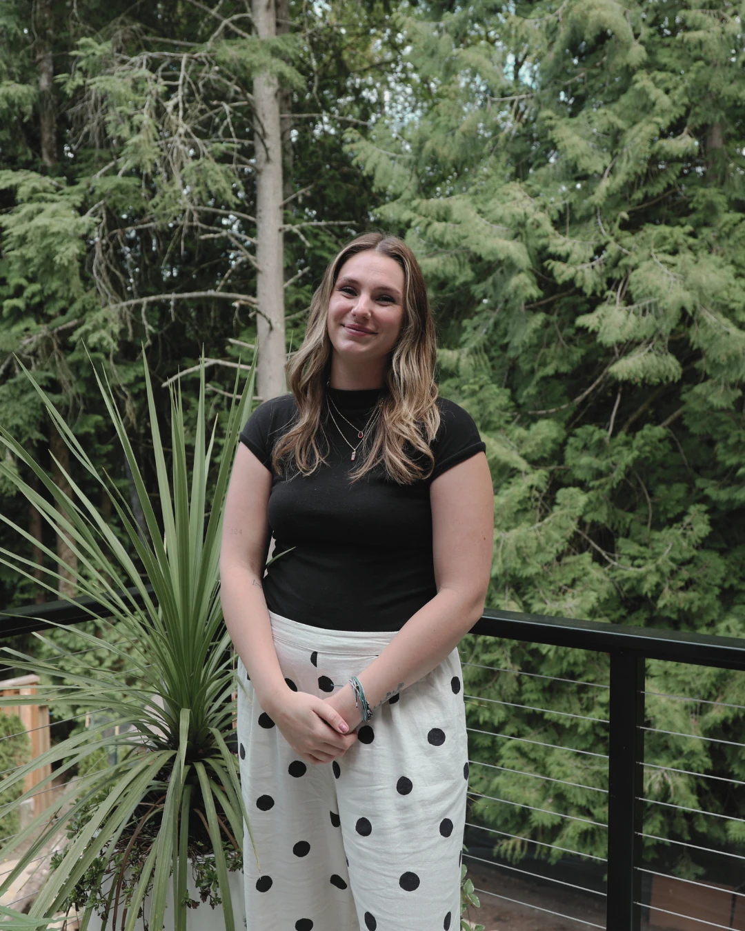 Sam, marketing director, standing outdoors on a balcony with greenery in the background.